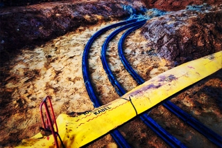 Offener Kabelgraben mit Dreierbuendeln dicker schwarzer Niederspannungskabel auf ockerfarbenem Sandbett - Projekt Produktionsanlage Solarlux - Offener Kabelgraben mit Dreierbuendeln dicker schwarzer Niederspannungskabel auf ockerfarbenem Sandbett, Kabel kommen oben rechts ins Bild machen 90-Grad Kurve und verlassen Bild unten rechts - Projekt Produktionsanlage Solarlux