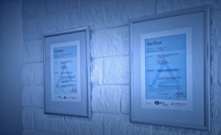 Zertifizierungen - in regelmässig stattfindenden Audits weisen wir die Erfüllung der Prüfungsstandards nach - Zertifikate Elmo Group für ISO 9001:2015 und SCC**2011 in Bilderrahmen an weißer Ziegel-Wand