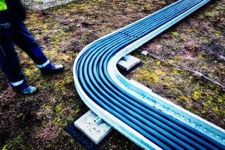 Kabeltrasse auf begruentem Dach kommt unten rechts ins Bild und laeuft rechts oben aus Bild heraus - Projekt Produktionsanlage Solarlux - Metallende Kabeltrasse auf begruentem Dach kommt unten rechts ins Bild, macht eine 90-Grad Abbiegung in Bildmitte und laeuft rechts oben aus Bild heraus - links davon sind Beine und Huefte eines Industrieelektrikers zu sehen - Projekt Produktionsanlage Solarlux